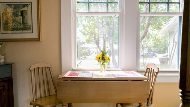 Una habitación con una mesa de madera y dos sillas, una ventana que da a árboles, y un cuadro en la pared.