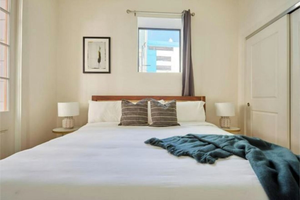 A hotel room with a large bed, two bedside tables with lamps, a window with curtains, and a door.
