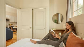 A room with a bed, a reading area, a kitchen in the background, and a large window.