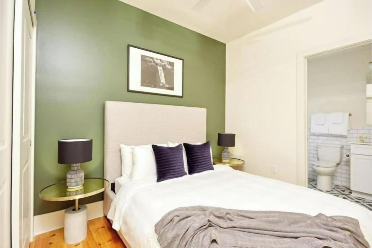 Une chambre à coucher avec un lit, deux tables de chevet, une commode, un tableau encadré au mur et une salle de bain visible à travers une porte ouverte.