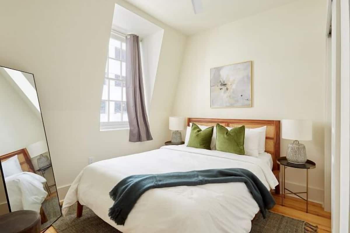 Quarto de hotel com uma cama grande, uma janela com cortinas, uma mesa de cabeceira com abajur e uma obra de arte na parede.