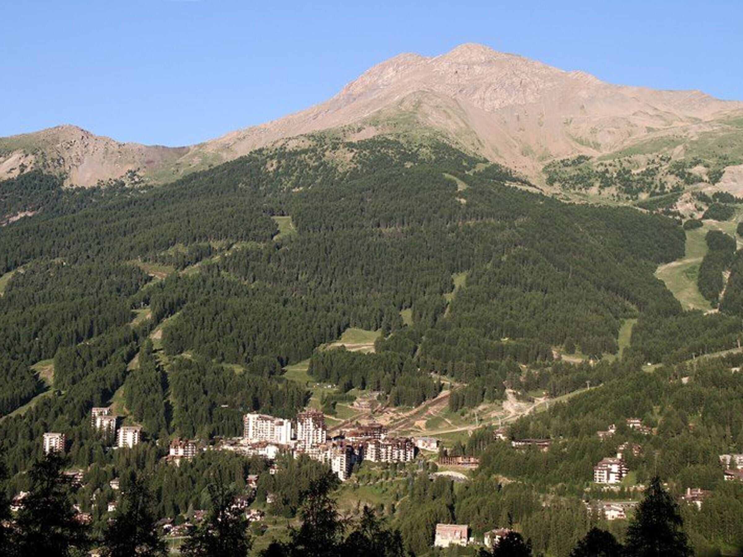 A mountainous area with dense forests and a cluster of buildings, likely a resort or hotel, with a clear sky.