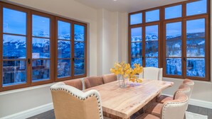A dining area with a large wooden table, cushioned chairs, and a view of snow-covered mountains through large windows. - Stunning 4-Bedroom Private Penthouse Villa With Panoramic Views (Mountain Village)