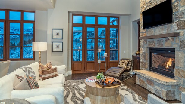 A spacious living room with a stone fireplace, large windows with a view of a snowy landscape, a comfortable seating area with a coffee table, and a television mounted on the wall. - Stunning 4-Bedroom Private Penthouse Villa With Panoramic Views (Mountain Village)