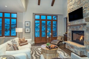 A spacious living room with a stone fireplace, large windows with a view of a snowy landscape, a comfortable seating area with a coffee table, and a television mounted on the wall. - Stunning 4-Bedroom Private Penthouse Villa With Panoramic Views (Mountain Village)