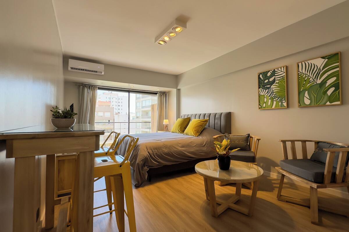 Una habitación de hotel moderna con una cama, un escritorio, una silla, una mesita y dos sillones. Hay dos ventanas amplias con cortinas, una lámpara de techo y dos obras de arte colgadas en la pared.