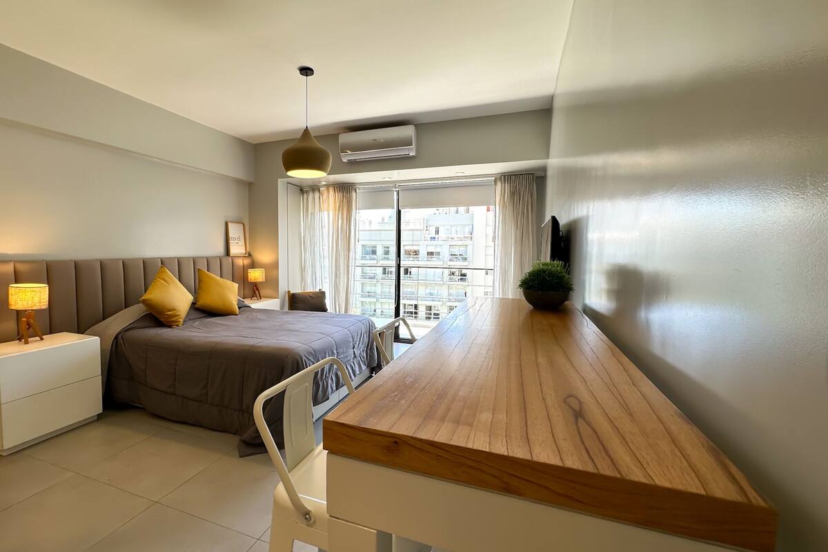 Un dormitorio moderno con cama, mesitas de noche, escritorio y vistas a la ciudad.
