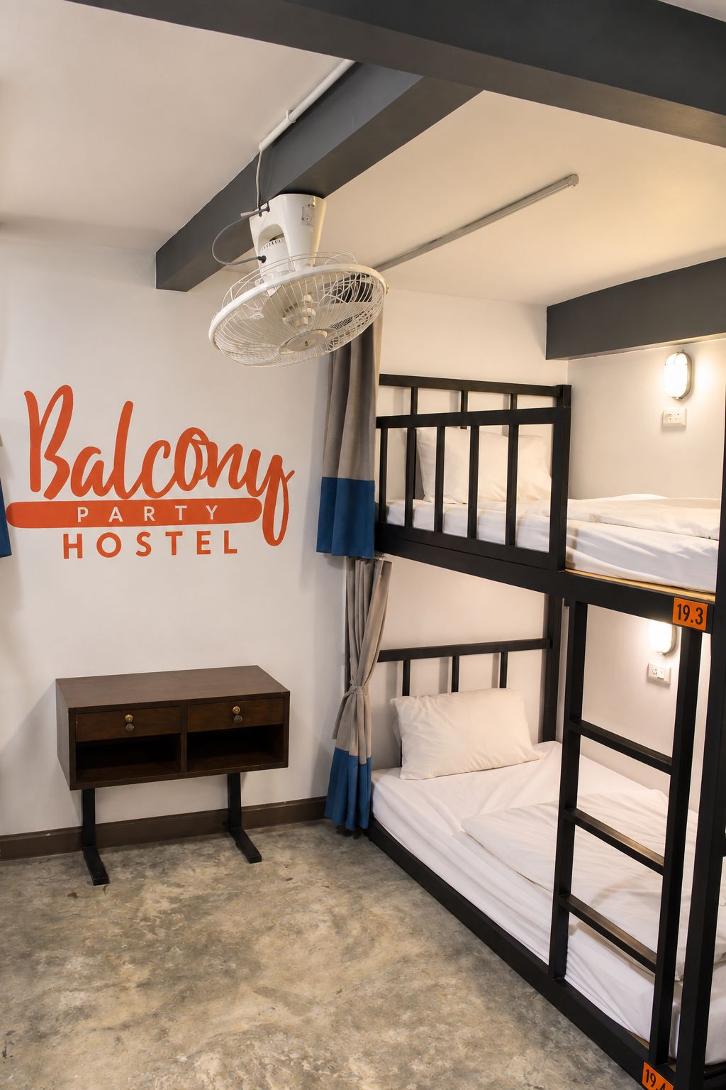 A hostel room with bunk beds, a ceiling fan, a bedside table, and a wall sign.