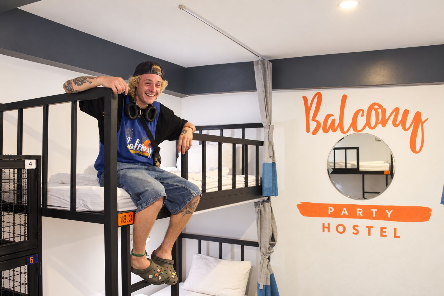 Una persona sentada en una cama individual en una habitación de albergue juvenil, con un cartel en la pared que dice 'Balcony PARTY HOSTEL'.