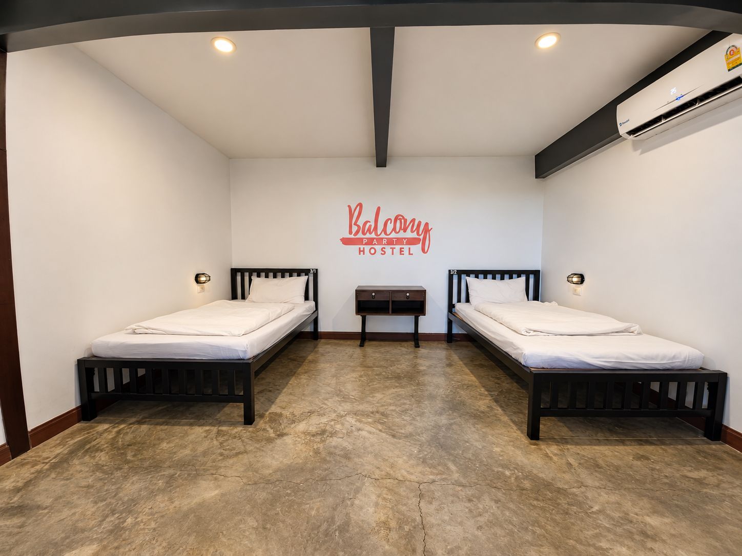 A hostel room with two beds, a small table, and a wall-mounted air conditioning unit.