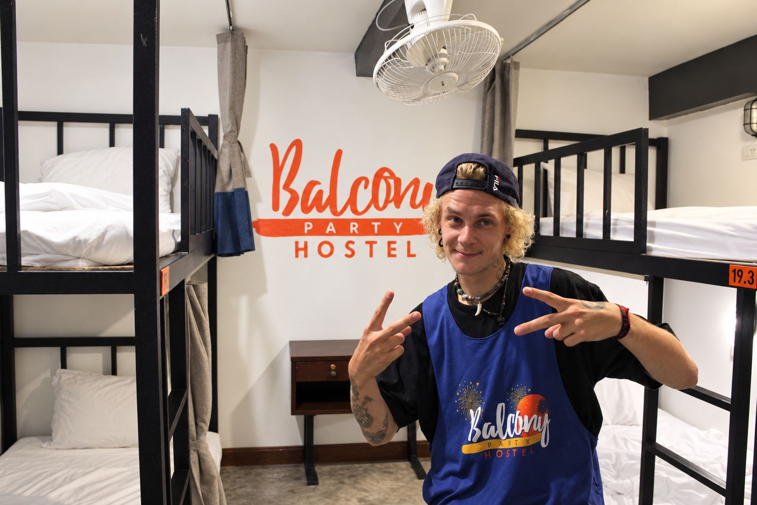 A bunk bed room with a sign reading 'Balcony Party Hostel' and a person wearing a 'Balcony Party Hostel' shirt.