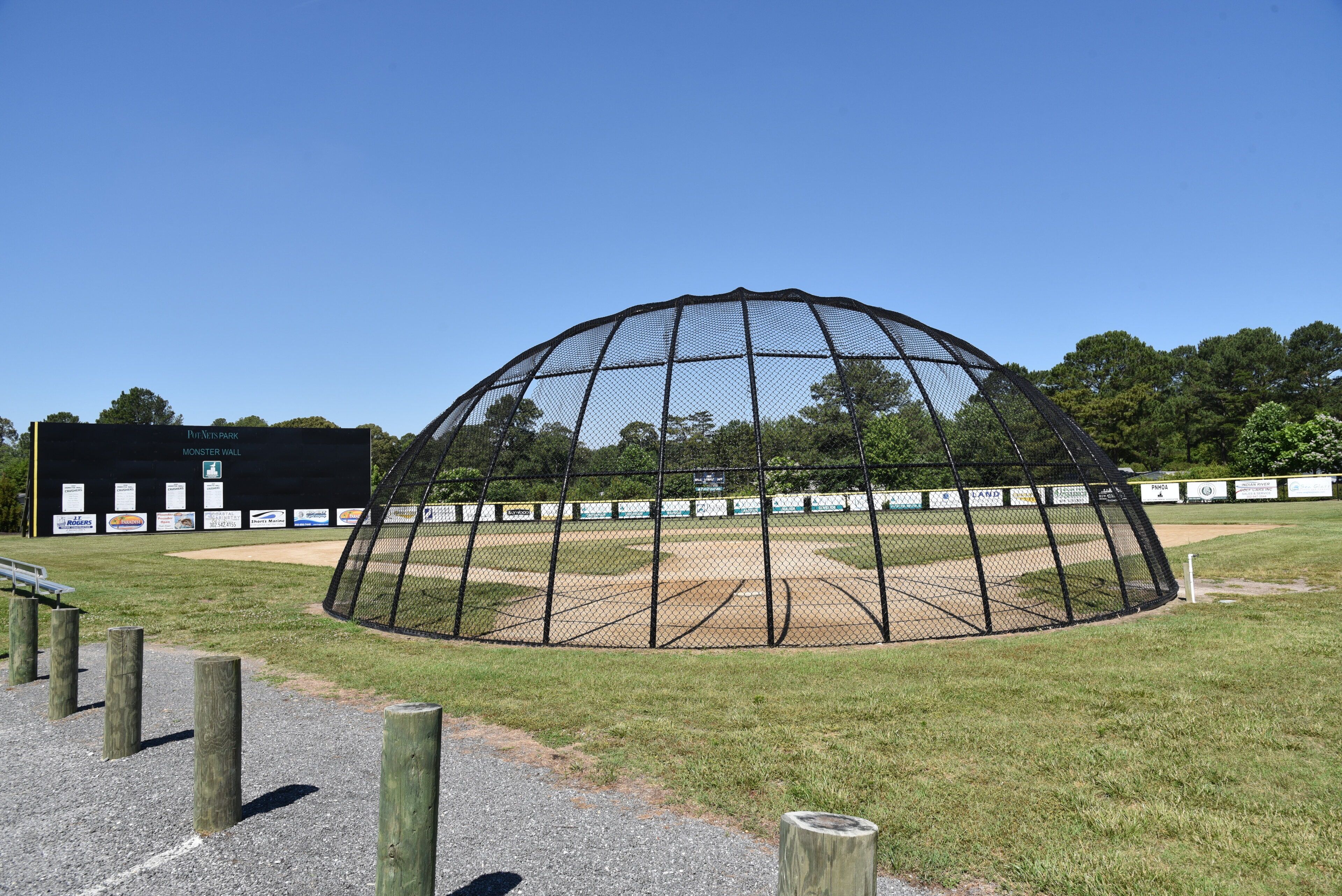 Un campo de béisbol con una jaula de bateo cubierta en malla, postes de madera y un edificio negro con el rótulo 'Monster Wall' al fondo.