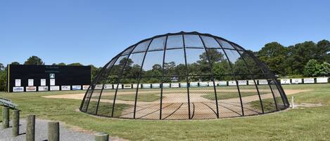 Un campo de béisbol con una jaula de bateo cubierta en malla, postes de madera y un edificio negro con el rótulo 'Monster Wall' al fondo.