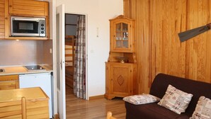 A compact living space with a kitchenette, wooden furniture, a sofa with patterned cushions, and a wooden cabinet.