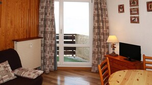 A cozy room with a wooden floor, a sofa with patterned cushions, a wooden table with a red cushion, a TV on a wooden stand, and a balcony with patterned curtains.