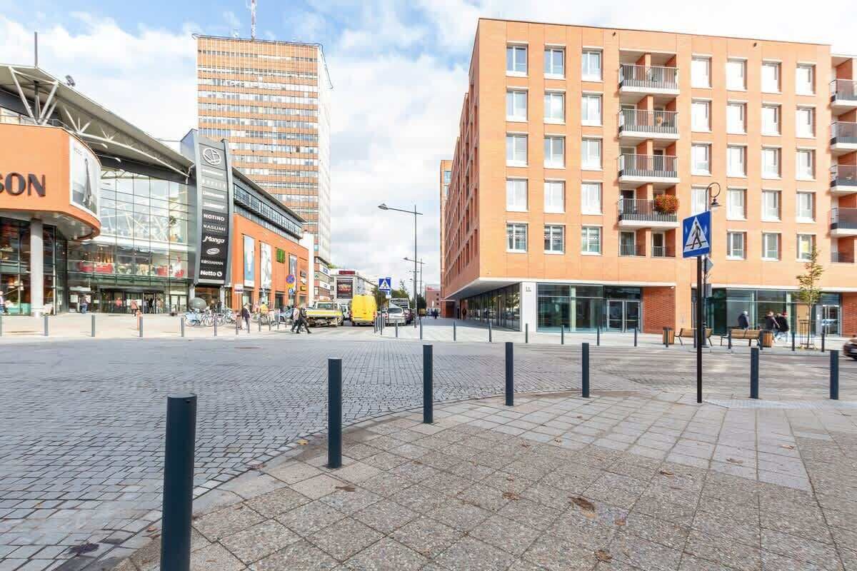 A modern urban square with a mix of commercial and residential buildings, including a prominent orange building with balconies and a glass-fronted structure.