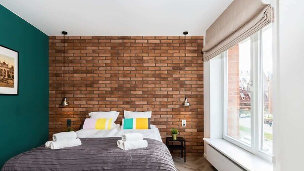 Una camera da letto con parete decorata con motivo a mattoni, un'ampia finestra, un letto con asciugamani ben piegati e due lampade a parete.