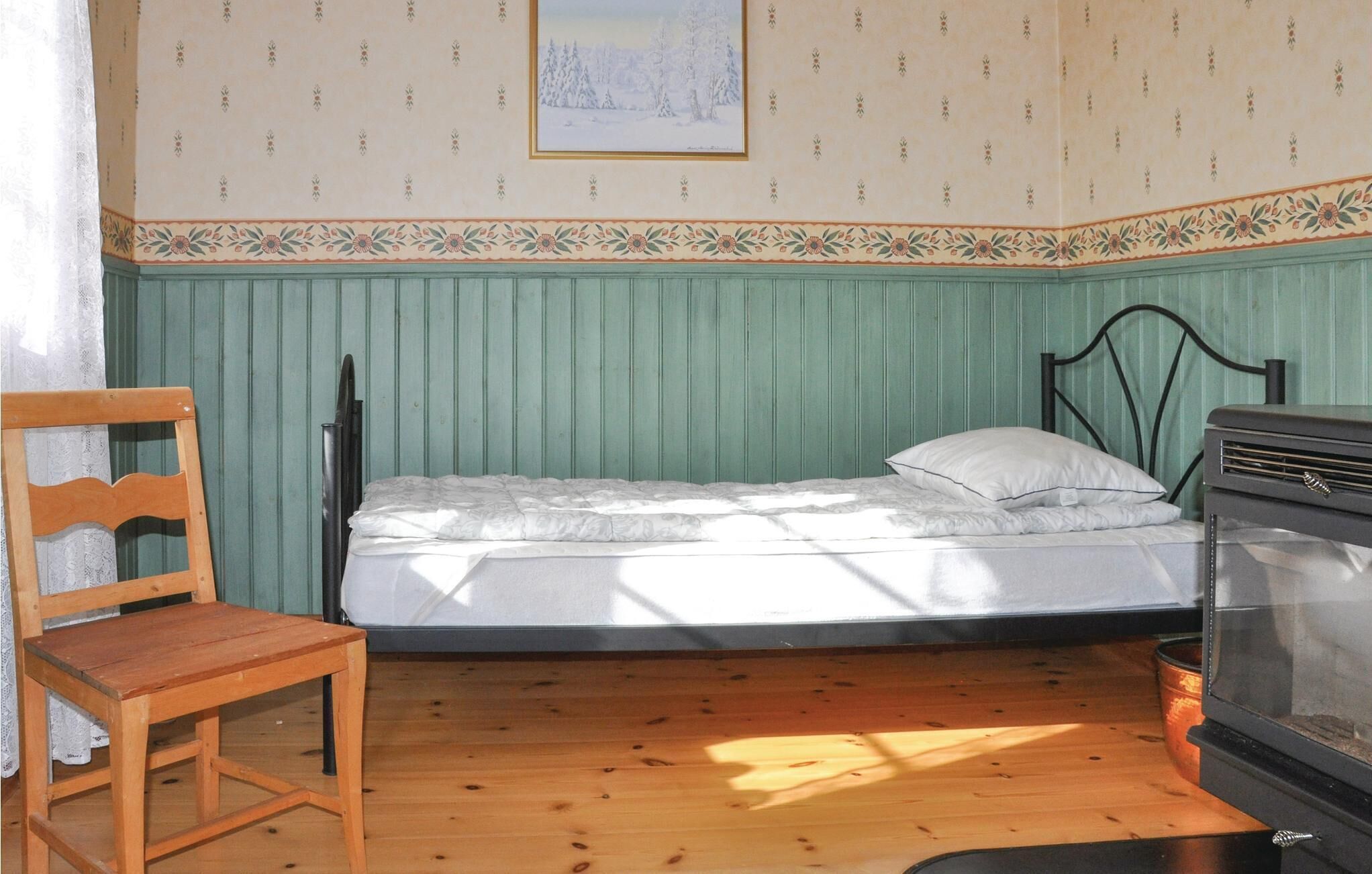 Quarto pequeno com uma cama de solteiro, uma cadeira de madeira, um radiador e um quadro na parede.