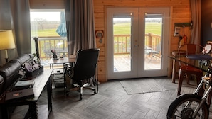 Een knus vertrek met houten inrichting, een zwarte bureaustoel, een eettafel, een motorfiets en uitzicht op een terras en gazon door de deur.
