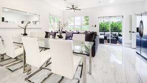A modern living room with a glass dining table, white chairs, a dark sofa, and a large window offering a view of greenery.