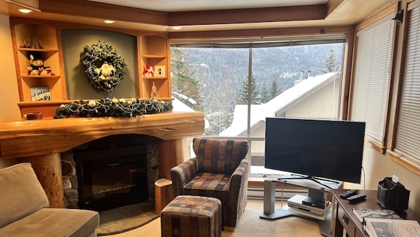 Un salon chaleureux avec une cheminée, un téléviseur et une vue sur des montagnes enneigées, visible à travers de grandes fenêtres.