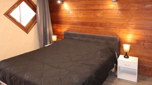 En pænt redt seng med et mørkt dynebetræk, et sengebord med en lampe og en trævæg med et vindue.