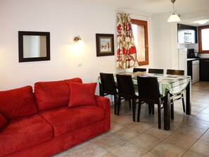 A living room with a red sofa, a dining table with chairs, a kitchen area with a microwave, and a window with curtains.
