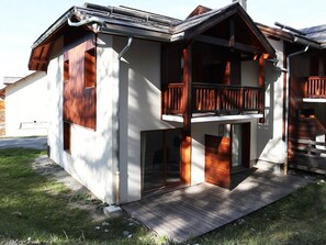 Una casa moderna de dos pisos con balcón de madera, ventanas grandes y un porche cubierto.