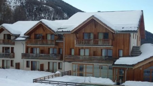 A multi-story wooden building with balconies, surrounded by snow-covered ground and mountains in the background. - Modern 2-Room Apt for 6 with Pool, Sauna, Hammam, Near Slopes (Les Orres)