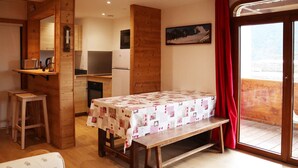 A cozy room with a wooden bench, a table with a quilt, a sofa, and a kitchen area with a refrigerator and microwave. - Ski-in/ski-out apartment for 6, balcony, near daycare (Les Orres)