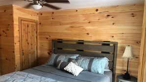 A wooden-paneled room with a bed, a ceiling fan, and a nightstand with a lamp.