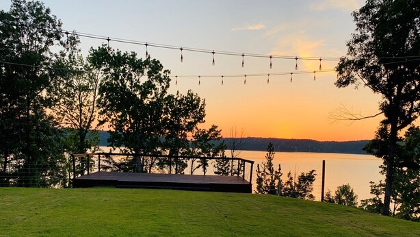 A grassy area with a wooden deck overlooking a lake, string lights, and trees.