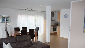 Een moderne woonkamer met een donkere bank, een eettafel met stoelen, een keuken met kasten en een klok aan de muur.