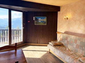 A room with a wooden floor, a sofa with patterned cushions, a wooden bench, and a large window with a mountain view.