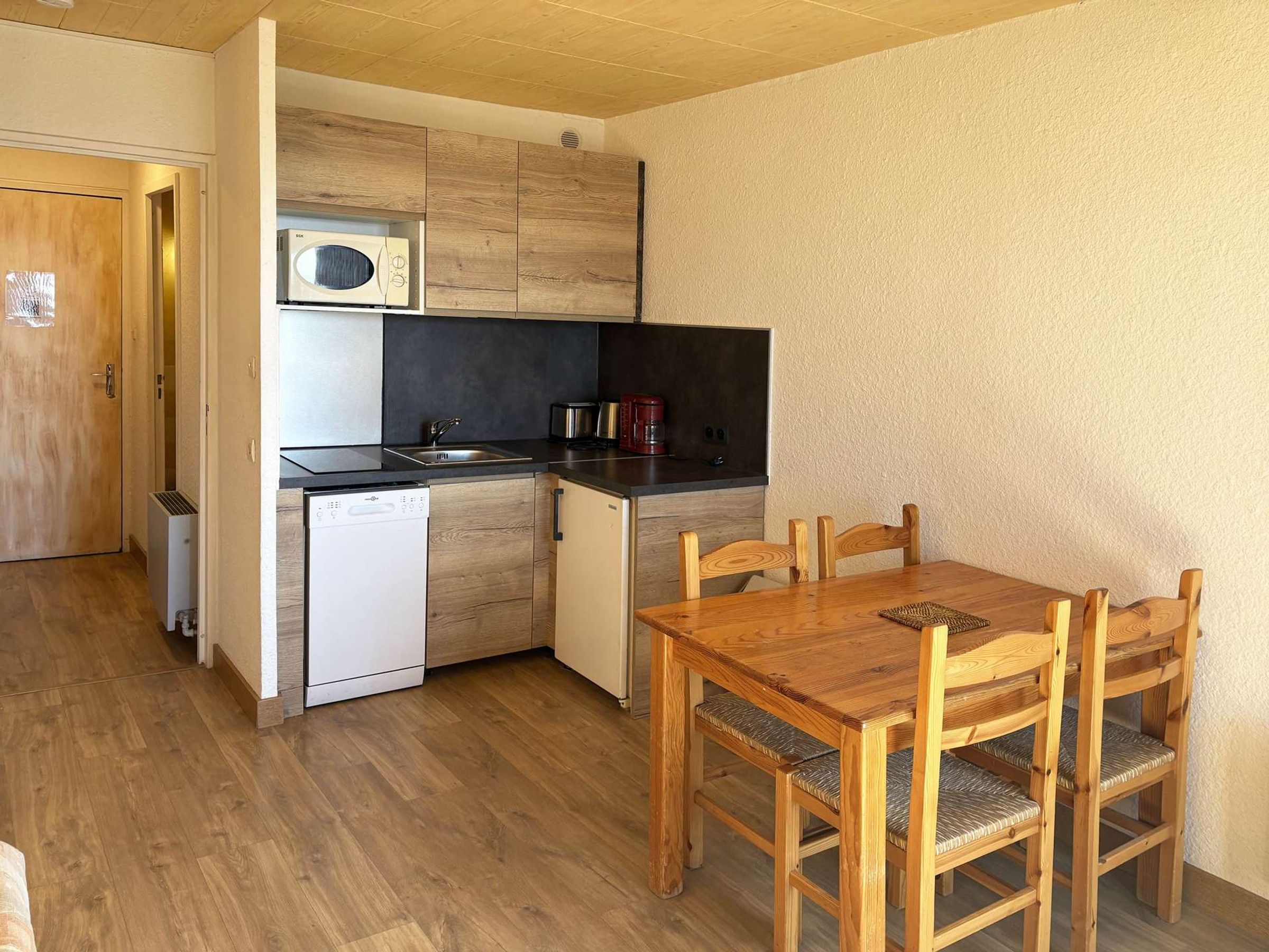 A compact kitchen with a dishwasher, a microwave, and a coffee maker. There is a wooden dining table with chairs.
