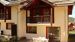 A two-story wooden house with a balcony, a patio with a table and chairs, and a view of the surrounding area. - Spacious Chalet for 10 with Fireplace, Ski-In/Ski-Out, Pool, Sauna & Parking (Les Orres)