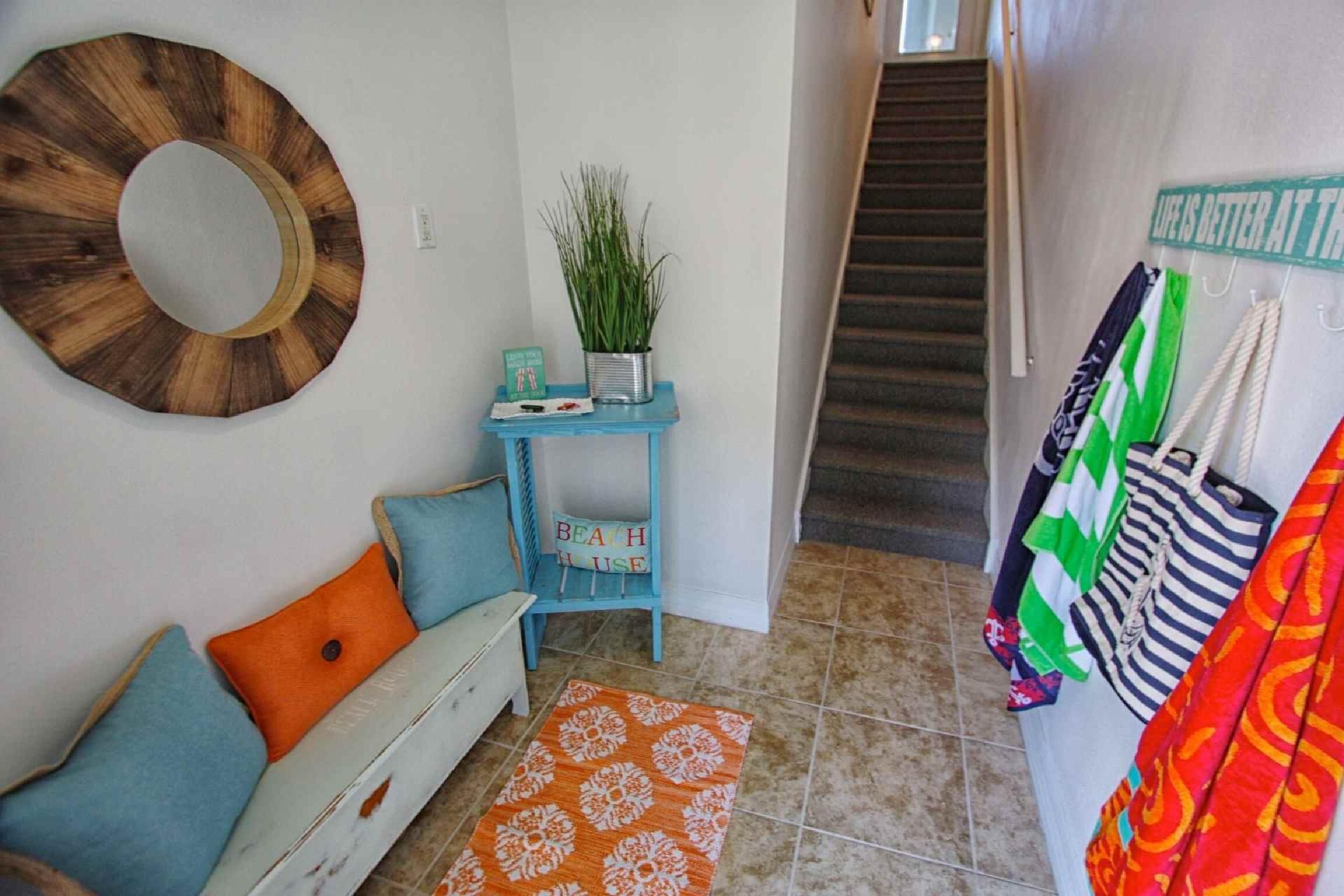 Un couloir avec un escalier, une chaise bleue, une petite table avec une plante et un tapis coloré.