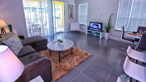 Uma sala de estar moderna com sofá, mesa de centro, televisão e porta de vidro deslizante que dá para uma varanda.