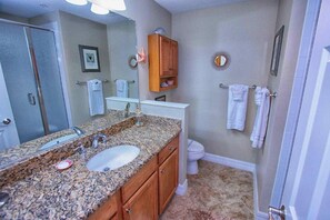 A bathroom with a granite countertop sink, wooden cabinets, a large mirror, a toilet, and a shower area.