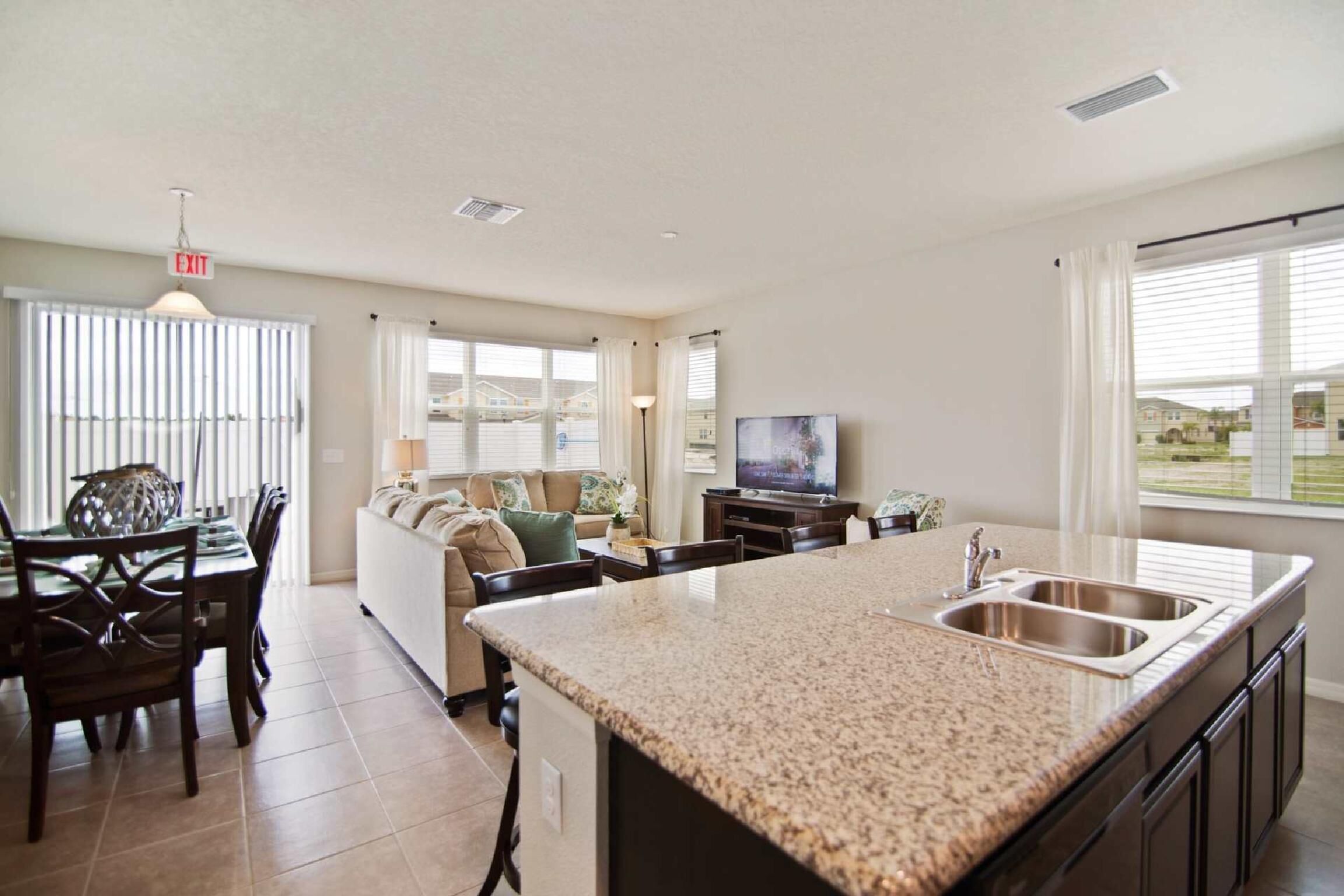 A modern kitchen with granite countertops, a dining area with a glass table, and a living room with a sofa and TV.