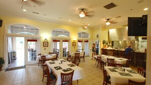 A dining area with multiple tables and chairs, a kitchen area, and a ceiling fan. - 1361 Tuscana (Davenport)