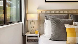 A neatly made bed with grey and white pillows, a bedside table with a lamp and a glass of orange juice, and a framed picture on the wall.