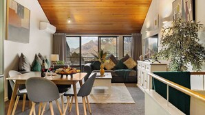 A modern living room with a wooden ceiling, a dining area with a table and chairs, a sofa with cushions, and a large window with a mountain view.