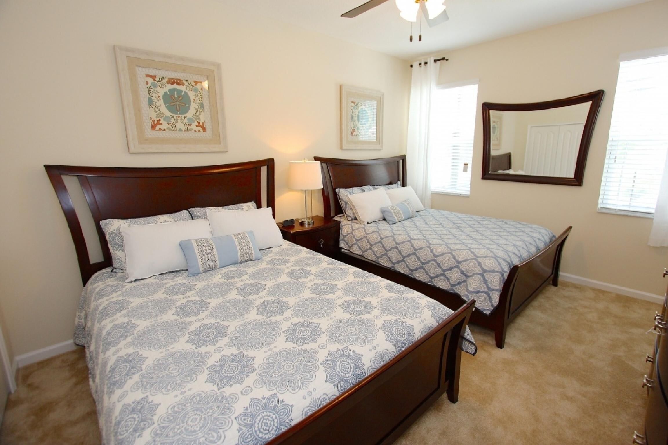 A bedroom with two beds, each with a wooden headboard and patterned bedding. There are two bedside tables with lamps, a ceiling fan, and a large mirror on the wall.