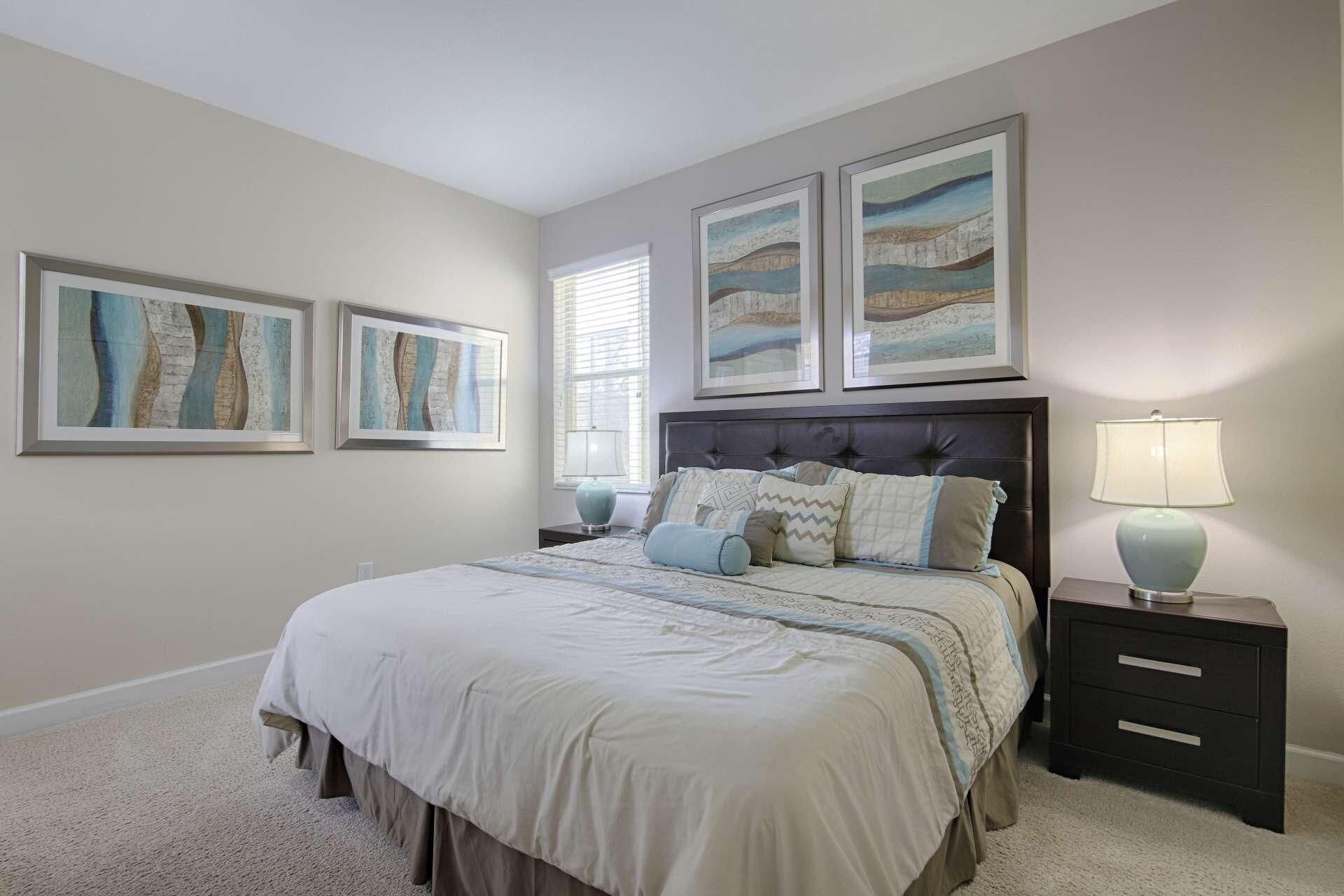 A bedroom with a large bed, a nightstand with a lamp, and three framed artworks on the wall.