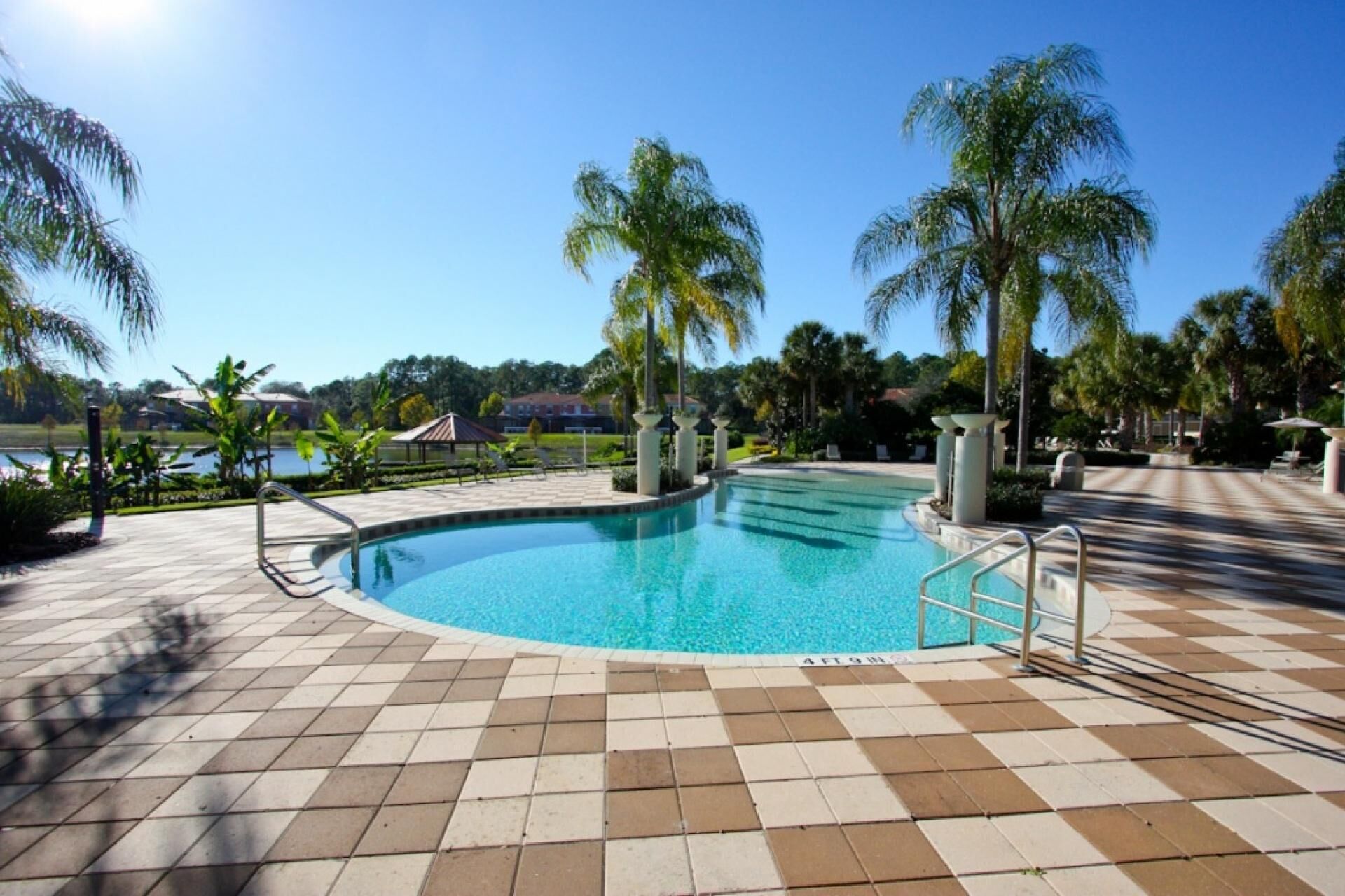 A swimming pool with tiled flooring and palm trees, surrounded by a landscaped area with a view of a lake and buildings in the background.