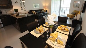 A modern kitchen with a central island, dining area with a black table and yellow napkins, and a living area with a black sofa and yellow armchair. - 17517 Dream (Clermont)