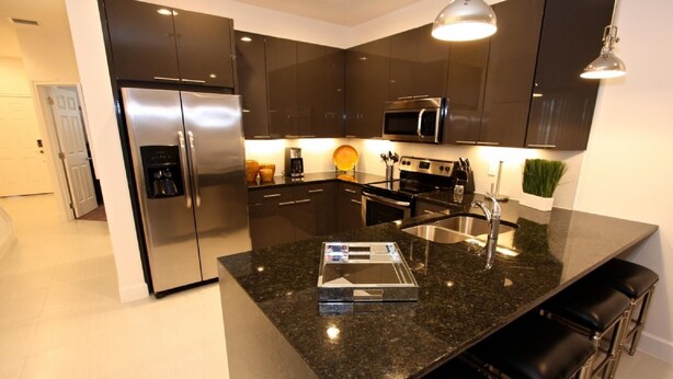A modern kitchen with dark cabinets, stainless steel appliances, a granite countertop, and bar stools. - 17517 Dream (Clermont)