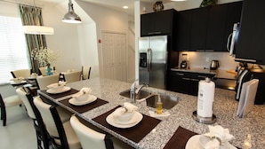 A modern kitchen with a granite countertop, stainless steel appliances, and a dining area with white chairs and a table set for four. - 17521 Dream (Clermont)
