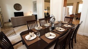 A dining area with a long table set for six, surrounded by wooden chairs. A sofa is visible in the background, and the room has a modern, clean design with a neutral color palette. - 4072 Solterra (Davenport)